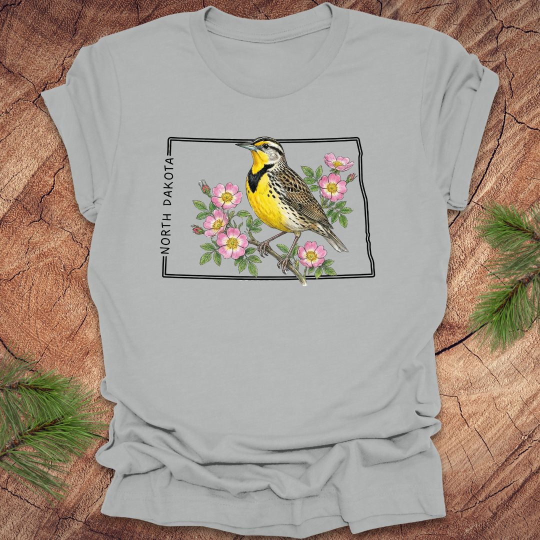 Silver t-shirt with a meadowlark and prairie rose flower inside the outline of North Dakota on a wooden surface