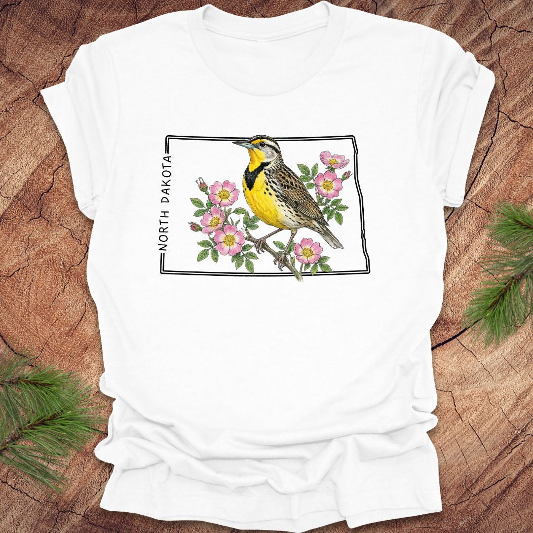 White t-shirt with a meadowlark and prairie rose flower inside the outline of North Dakota on a wooden surface