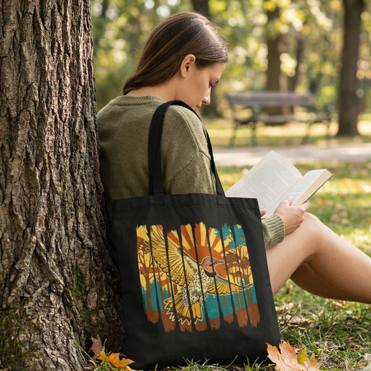 Woman reading a book in a park with a black tote bag featuring a colorful bird design.