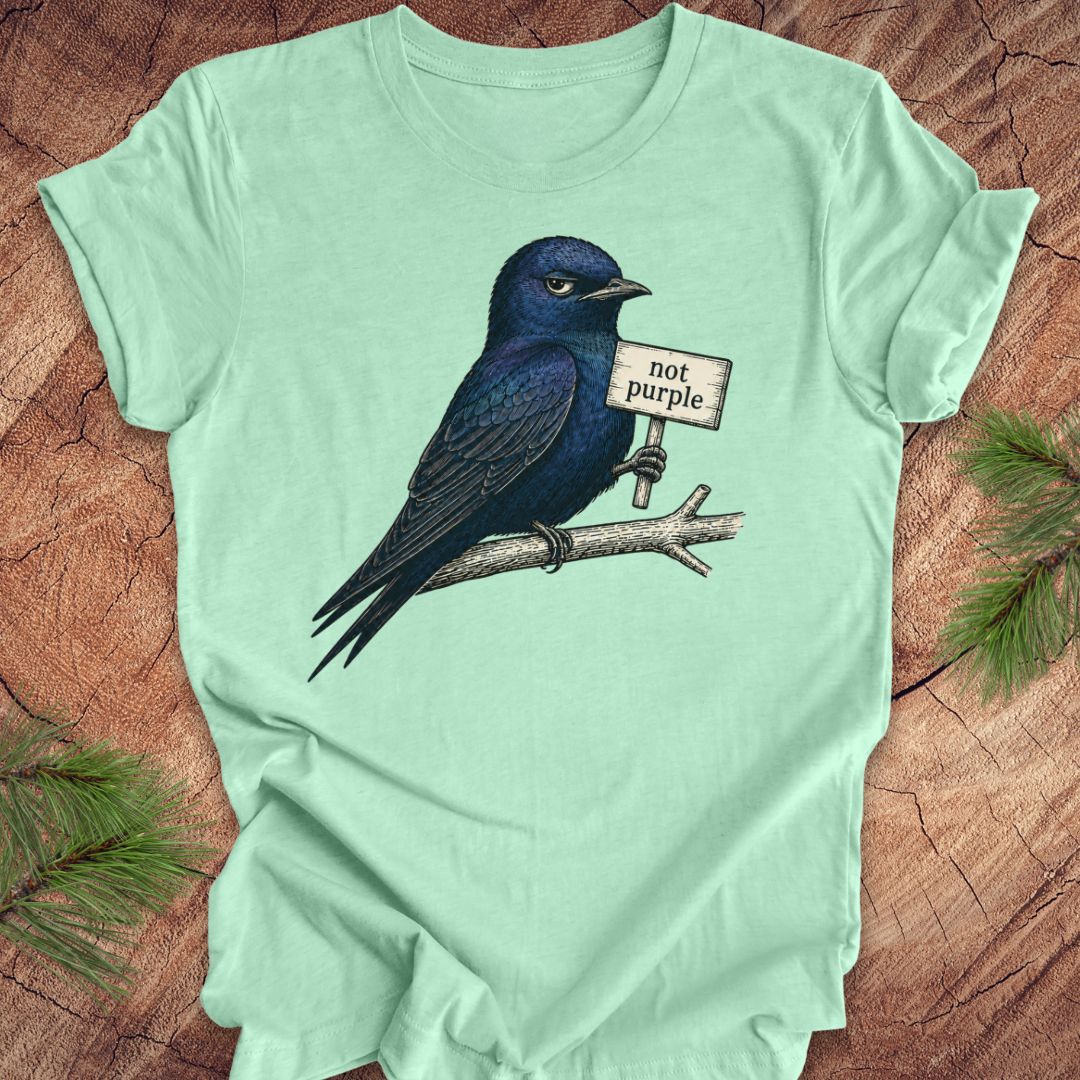 Mint green t-shirt with a purple martin bird holding a 'not purple' sign on a wooden surface.