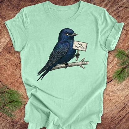 Mint green t-shirt with a purple martin bird holding a 'not purple' sign on a wooden surface.