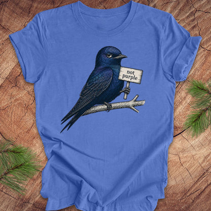 Blue t-shirt with a bird holding a 'not purple' sign on a wooden surface.