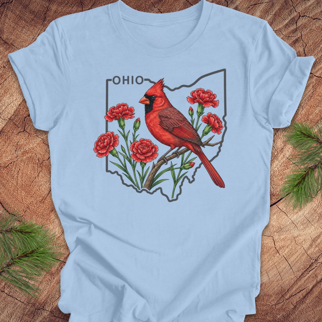 A light blue short-sleeve t-shirt laid flat on a rustic wood surface, featuring a graphic of a red cardinal perched on flowers inside the outline of Ohio with the word 'OHIO' above it.