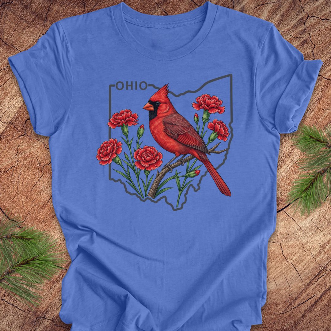 A blue short-sleeve crewneck t-shirt laid flat on a rustic wood background with pine branches in the corners. The front of the shirt features a graphic of a red cardinal perched on red carnations inside the outline of the state of Ohio, with the word 'OHIO' printed above.