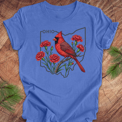 A blue short-sleeve crewneck t-shirt laid flat on a rustic wood background with pine branches in the corners. The front of the shirt features a graphic of a red cardinal perched on red carnations inside the outline of the state of Ohio, with the word 'OHIO' printed above.