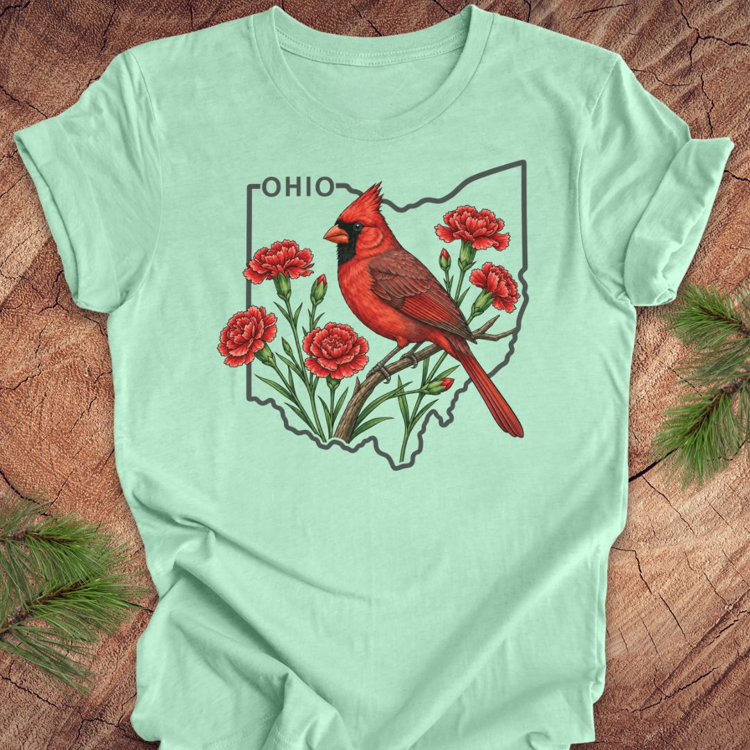 A mint green short-sleeve t-shirt laid flat on a rustic wood background with pine branches in the corners. The shirt features a graphic print of a red cardinal perched on a branch with red flowers inside the outline of Ohio, with the word 'OHIO' printed above.