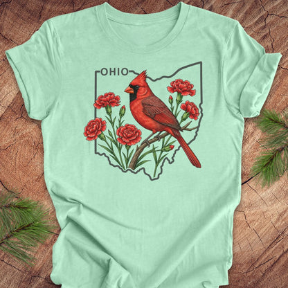 A mint green short-sleeve t-shirt laid flat on a rustic wood background with pine branches in the corners. The shirt features a graphic print of a red cardinal perched on a branch with red flowers inside the outline of Ohio, with the word 'OHIO' printed above.