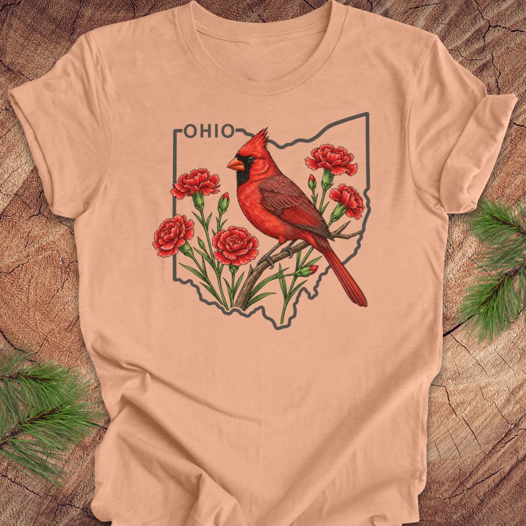 A peach-colored short-sleeve t-shirt laid flat on a rustic wood background, featuring a graphic of a red cardinal perched on flowers inside the outline of Ohio with the word 'OHIO' above.