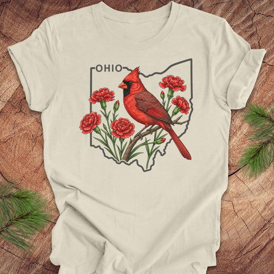 An off-white t-shirt laid flat on a rustic wood background, featuring a graphic of a red cardinal perched on a branch with red flowers inside the outline of the state of Ohio, with the word 'OHIO' printed above.
