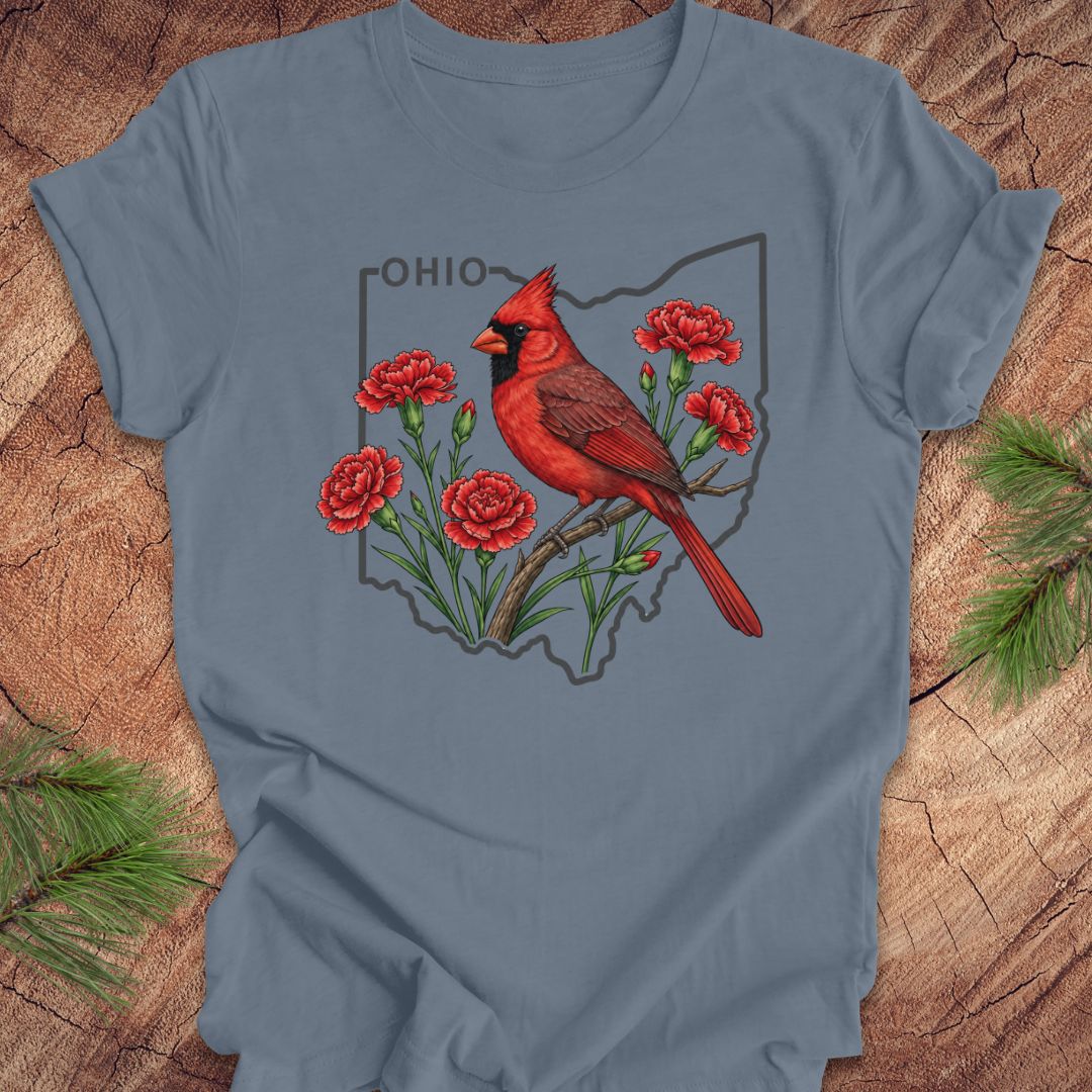 A slate blue crewneck t-shirt laid flat on a rustic wood background with pine needles in the corners. The shirt features a graphic print of a red cardinal bird perched on a branch with red flowers, set against an outline of the state of Ohio and the word 'OHIO'.