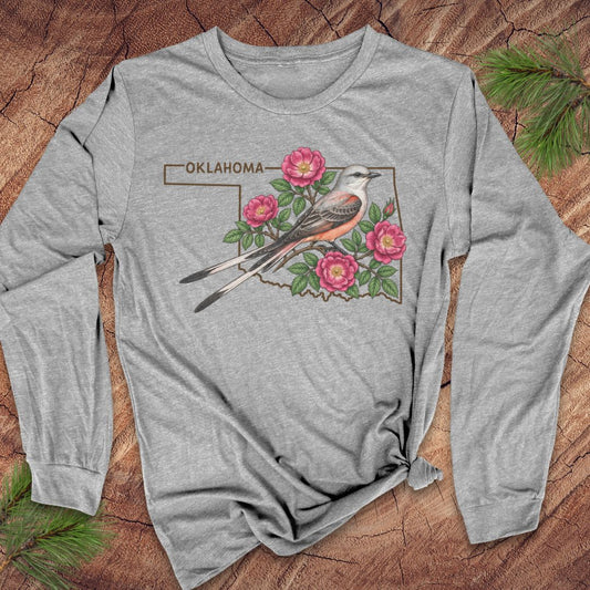 A heather gray long-sleeve t-shirt with a graphic of an Oklahoma state outline, a king fly bird, and pink flowers, laid flat on a rustic wood background with pine needles.