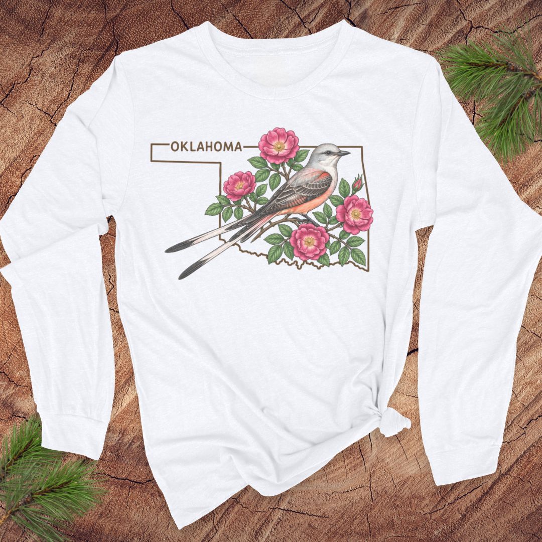 A white long-sleeve t-shirt with a graphic of a bird, pink flowers, and the word 'OKLAHOMA' printed on the front, styled flat on a rustic wood background with pine branches.