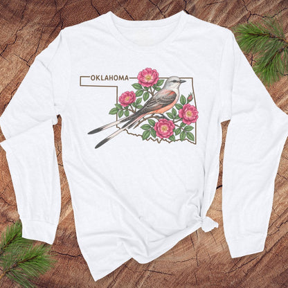 A white long-sleeve t-shirt with a graphic of a bird, pink flowers, and the word 'OKLAHOMA' printed on the front, styled flat on a rustic wood background with pine branches.