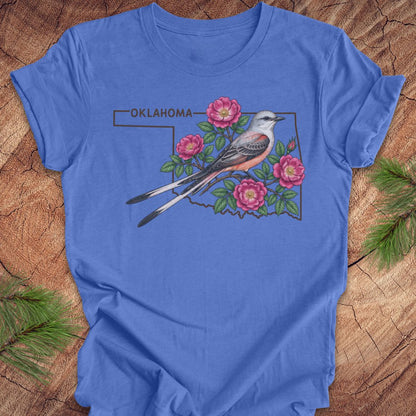 A royal blue short-sleeve t-shirt laid flat on a rustic wood background with pine branches in the corners. The shirt features a graphic of an Oklahoma state outline with a gray and orange bird perched on pink flowers and green leaves, with the word 'OKLAHOMA' above.