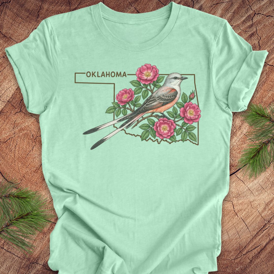 A mint green short-sleeve t-shirt laid flat on a rustic wood background with pine branches in the corners. The shirt features a graphic of an Oklahoma state outline with a Western Scuppers bird perched on pink roses and the word 'OKLAHOMA' above.