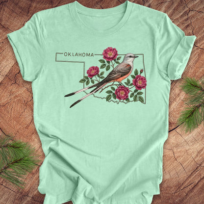 Mint green t-shirt with state bird, scissor-tailed flycatcher and rose, on an Oklahoma map, placed on a wooden surface.