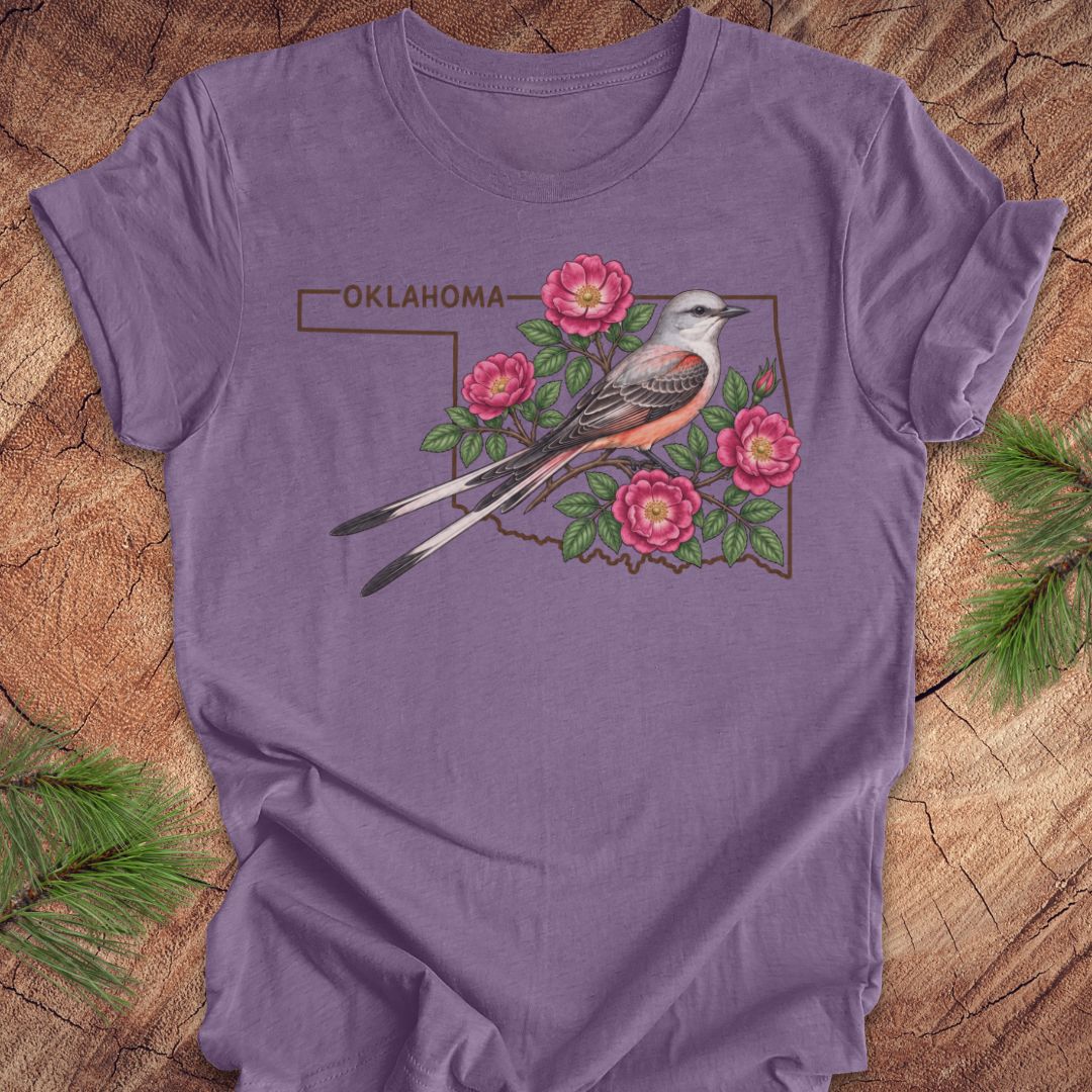 A purple short-sleeve t-shirt laid flat on a rustic wood background with pine branches in the corners. The shirt features a graphic of the state outline of Oklahoma with a gray and orange bird, pink flowers, and the word 'OKLAHOMA' printed above.