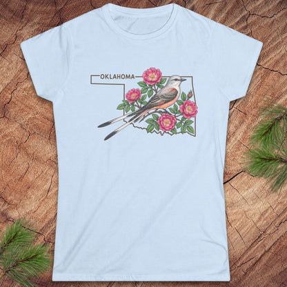 A light blue short-sleeve crewneck t-shirt featuring a graphic print of an Oklahoma state outline with a bird, pink flowers, and the word 'OKLAHOMA' at the top, displayed flat on a rustic wood background with pine branches.