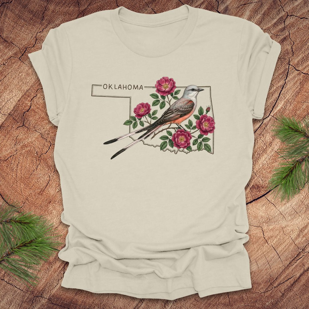 Beige t-shirt with state bird, scissor-tailed flycatcher and rose, on an Oklahoma map, placed on a wooden surface.