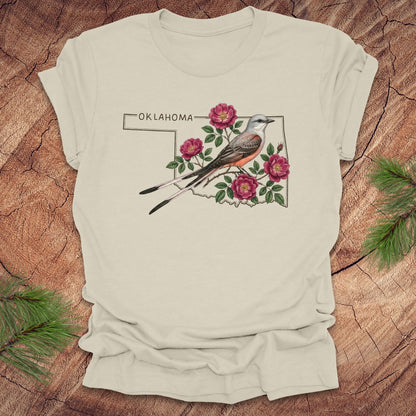 Beige t-shirt with state bird, scissor-tailed flycatcher and rose, on an Oklahoma map, placed on a wooden surface.