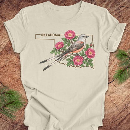 An off-white t-shirt laid flat on a rustic wood background, featuring a graphic of a Western Scott's oriole bird perched on pink roses inside the outline of Oklahoma, with the word 'OKLAHOMA' above.