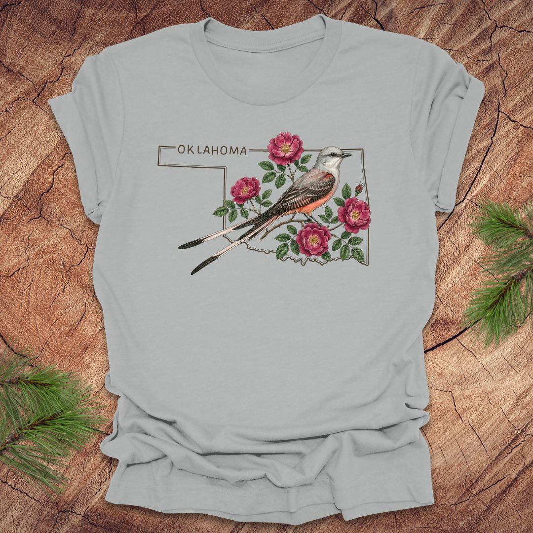 Silver t-shirt with state bird, scissor-tailed flycatcher and rose, on an Oklahoma map, placed on a wooden surface.