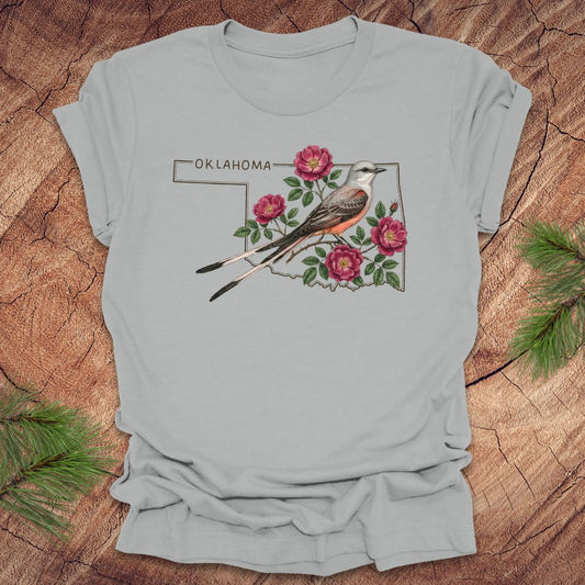 Silver t-shirt with state bird, scissor-tailed flycatcher and rose, on an Oklahoma map, placed on a wooden surface.