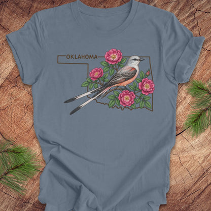 A slate blue short-sleeve t-shirt laid flat on a rustic wood background with pine branches in the corners. The shirt features a graphic of an Oklahoma state outline containing pink flowers, green leaves, and a Western Red-shouldered Flycatcher with 'OKLAHOMA' text above.