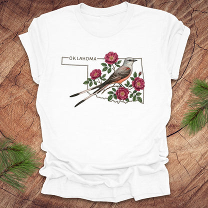 White t-shirt with state bird, scissor-tailed flycatcher and rose, on an Oklahoma map, placed on a wooden surface.