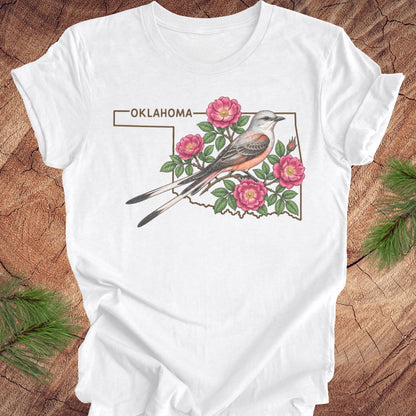 A white short-sleeve T-shirt laid flat on a rustic wood surface, featuring a printed Oklahoma state outline graphic with a bird and pink flowers. Pine branches are visible at the corners of the frame.