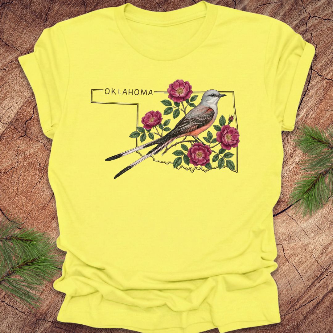 Yellow t-shirt with a graphic of a bird and flowers on an outline of Oklahoma, placed on a wooden surface.