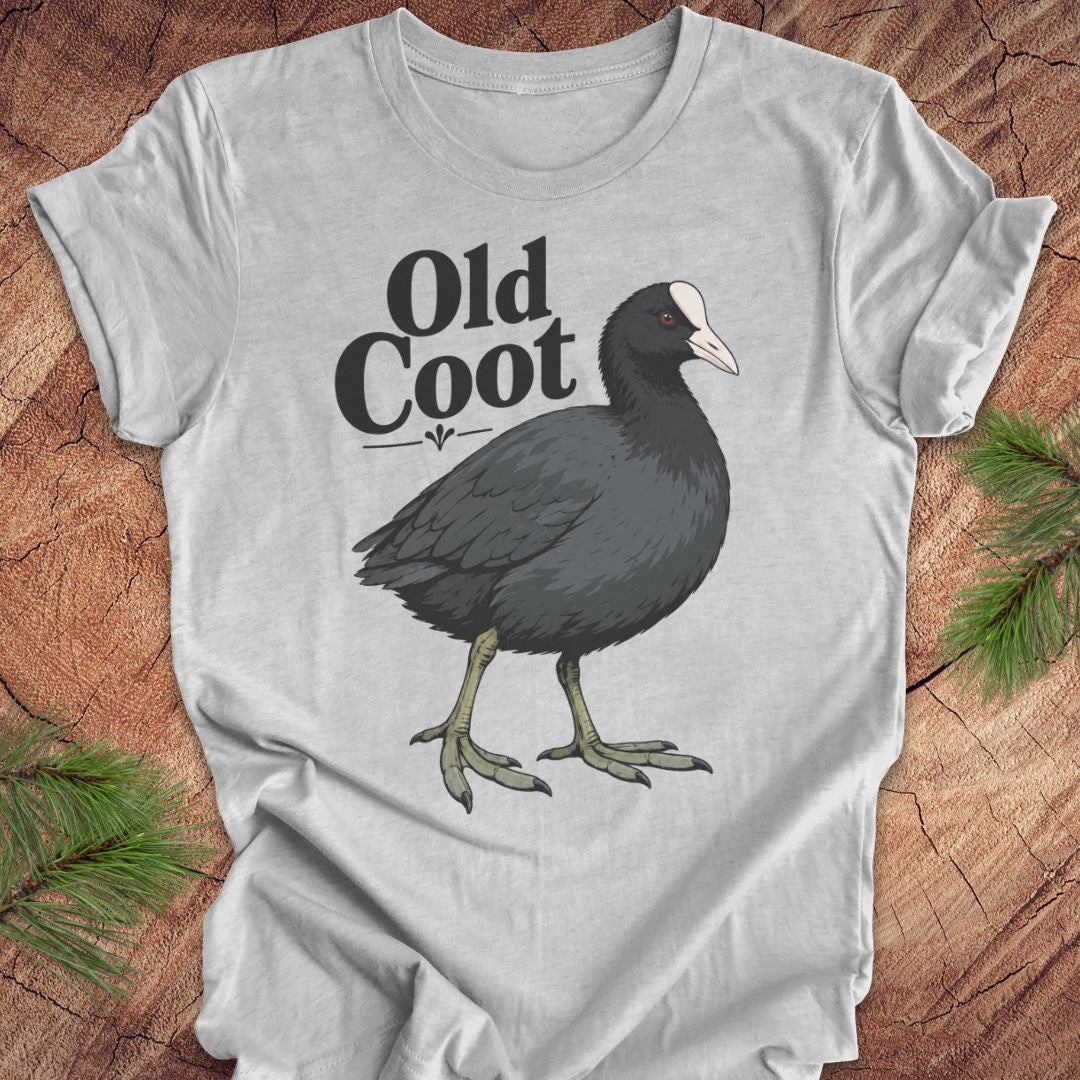 A light gray short-sleeve t-shirt laid flat on a rustic wood background with pine branches in the corners. The shirt features a graphic of a dark gray coot with a white beak and the text 'Old Coot' printed above it.