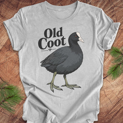 A light gray short-sleeve t-shirt laid flat on a rustic wood background with pine branches in the corners. The shirt features a graphic of a dark gray coot with a white beak and the text 'Old Coot' printed above it.
