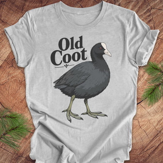 A light gray short-sleeve t-shirt laid flat on a rustic wood background with pine branches in the corners. The shirt features a graphic of a dark gray coot with a white beak and the text 'Old Coot' printed above it.