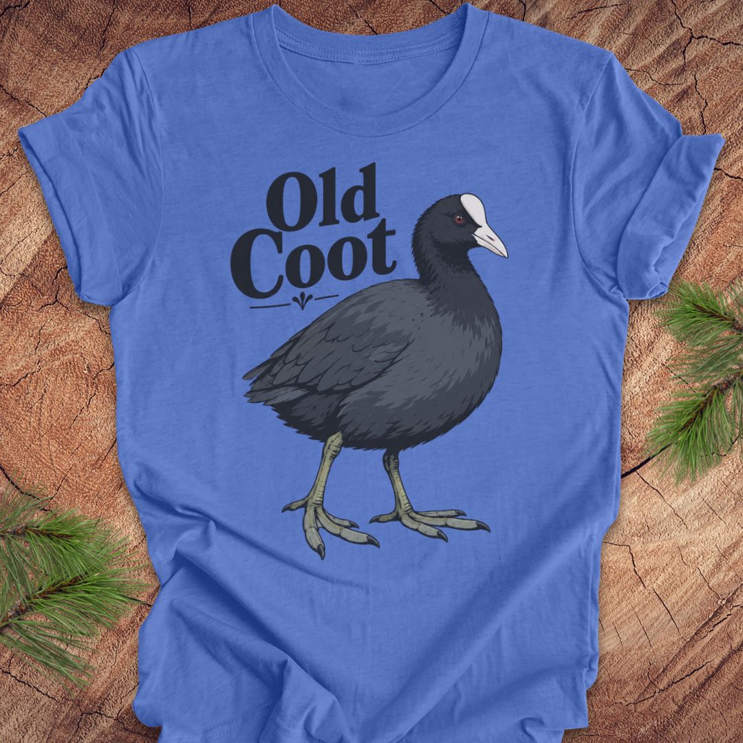 A blue short-sleeve t-shirt laid flat on a rustic wood surface, featuring a printed illustration of a dark coot bird and the words 'Old Coot' in black. Pine branches are visible at the corners of the frame.