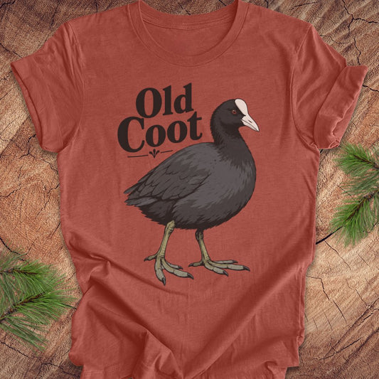 A rust-orange crewneck t-shirt featuring a graphic of an 'Old Coot' bird with black text, laid flat on a rustic wood background with pine needles.