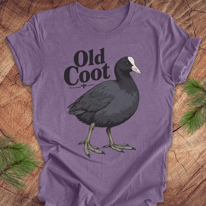 A purple short-sleeve t-shirt laid flat on a rustic wood background with pine branches in the corners. The shirt features a graphic of a dark gray coot bird with a white beak and the text 'Old Coot' in black.