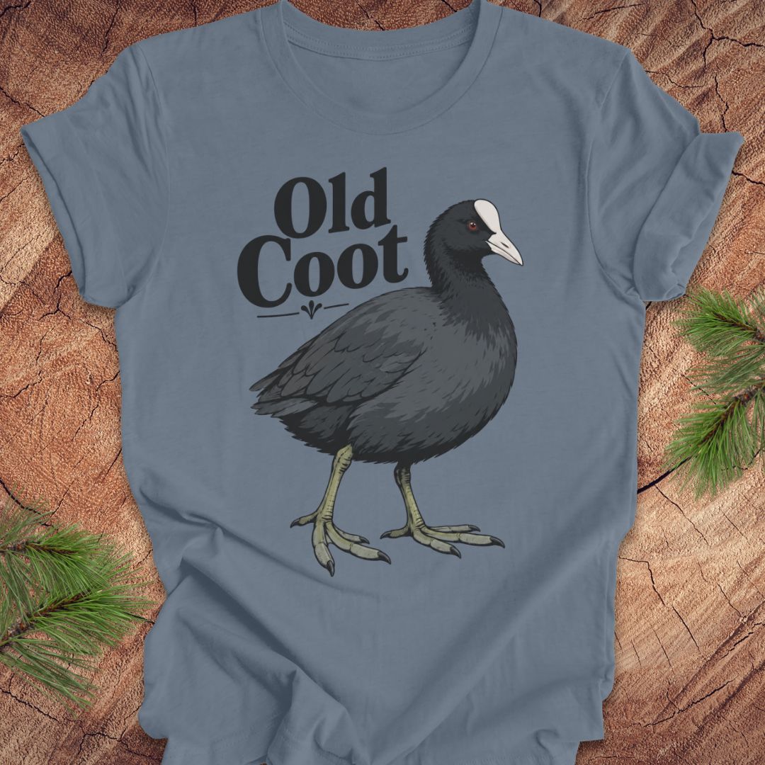 A slate blue t-shirt featuring a graphic of an American coot bird and the text 'Old Coot' in black, laid flat on a rustic wood background with pine branches in the corners.