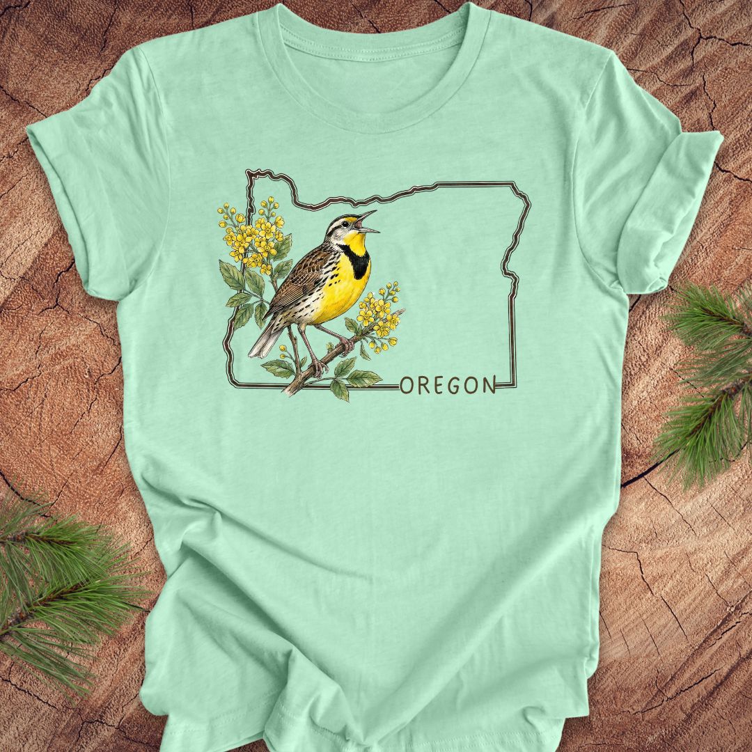 Mint green t-shirt with a bird and floral design on an outline of Oregon, placed on a wooden surface.