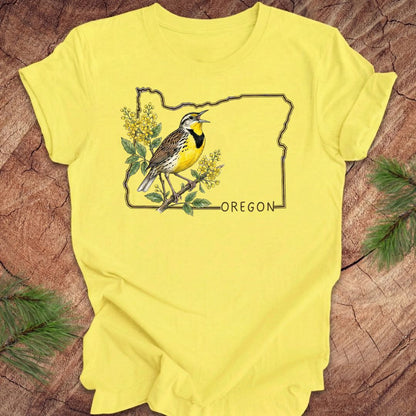Yellow t-shirt with a bird and floral design on an outline of Oregon, placed on a wooden surface.