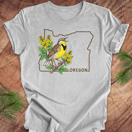 A light gray short-sleeve t-shirt laid flat on a rustic wood background with pine branches in the corners. The shirt features a graphic of a yellow and brown bird perched on a branch with yellow flowers, inside an outline of Oregon with the word 'OREGON' printed below.