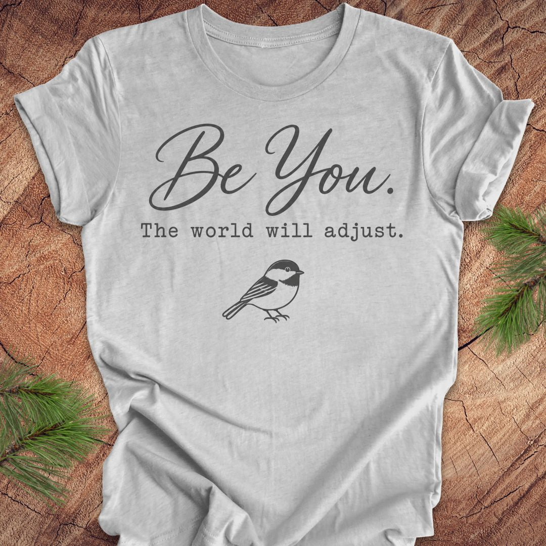 A light gray graphic t-shirt laid flat on a rustic wood background with pine branches in the corners. The shirt features the text 'Be You.