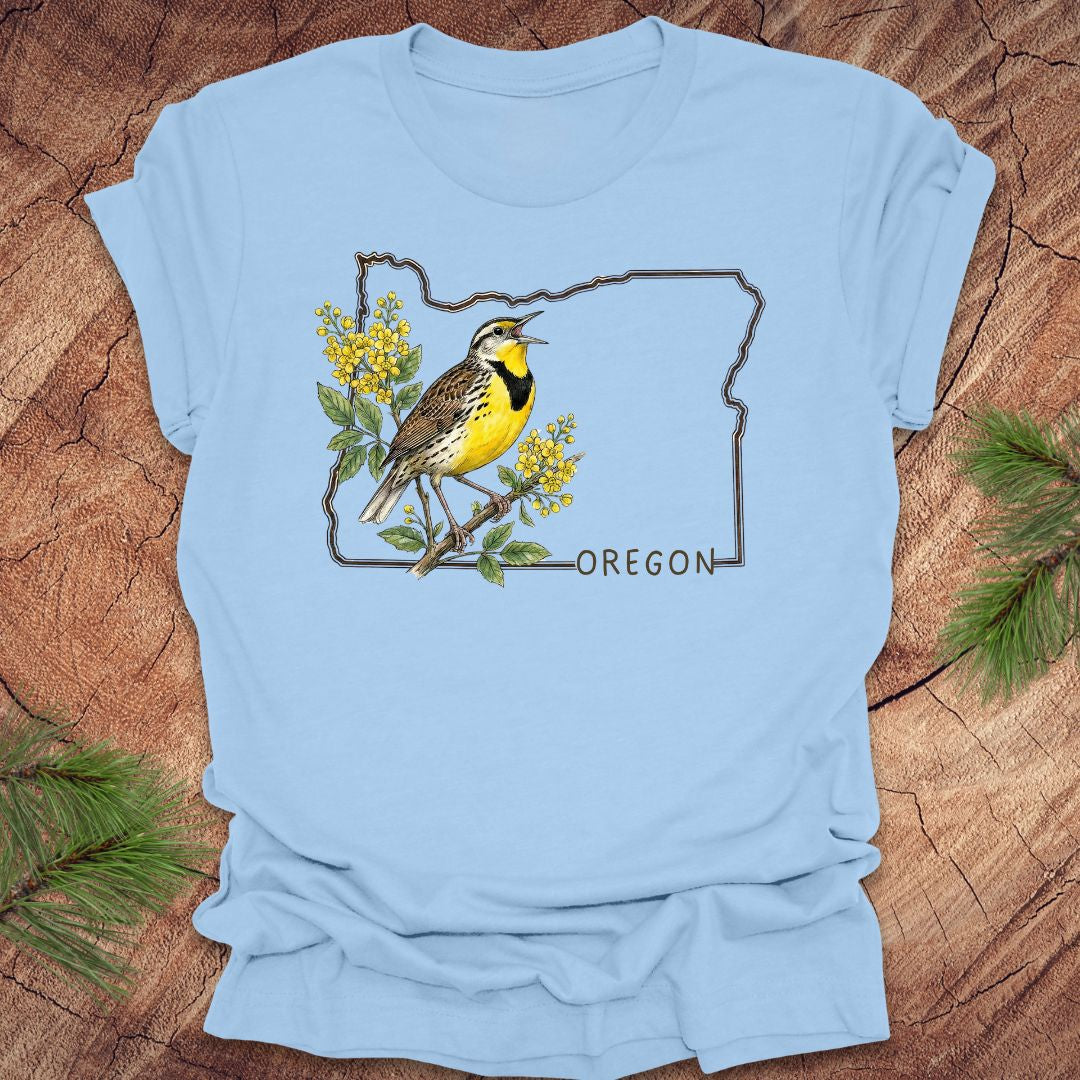 Baby blue t-shirt with a bird and floral design on an outline of Oregon, placed on a wooden surface.