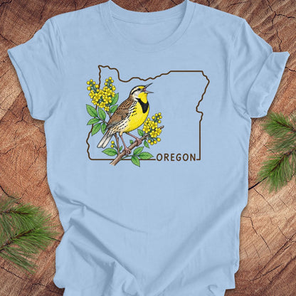 A light blue short-sleeve crewneck t-shirt laid flat on a rustic wood background with pine needles. The shirt features a graphic of a yellow and brown bird inside the outline of Oregon, with yellow flowers and the word 'OREGON' printed below.