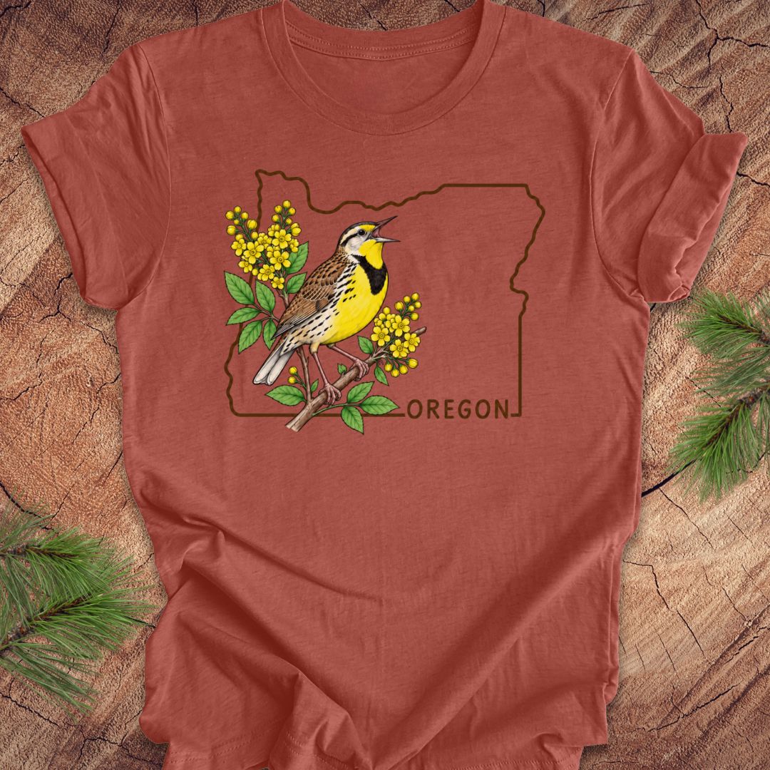 A rust-colored short-sleeve t-shirt laid flat on a rustic wood background, featuring a graphic of an Oregon state outline with a yellow bird and yellow flowers, and the word 'OREGON' printed below.