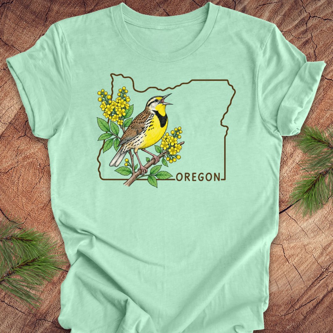 A mint green short-sleeve t-shirt laid flat on a rustic wood background, featuring a graphic of an Oregon state outline with a yellow-breasted bird, green leaves, yellow flowers, and the word 'OREGON'.