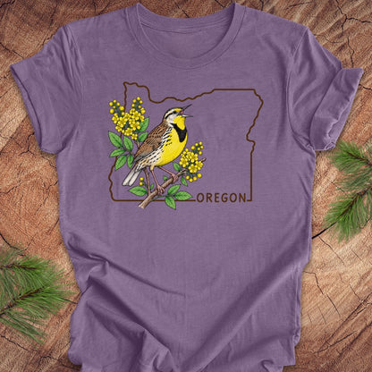 A purple short-sleeve t-shirt laid flat on a rustic wood background with pine branches in the corners. The shirt features a graphic of a yellow and brown bird perched on a branch with yellow flowers, outlined by the state shape of Oregon and the word 'OREGON'.