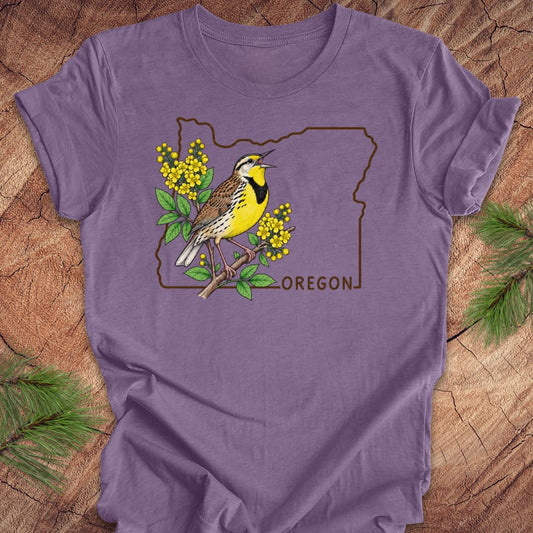A purple short-sleeve t-shirt laid flat on a rustic wood background with pine branches in the corners. The shirt features a graphic of a yellow and brown bird perched on a branch with yellow flowers, outlined by the state shape of Oregon and the word 'OREGON'.