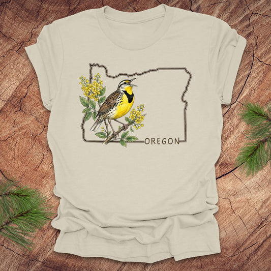 Beige t-shirt with a bird and floral design on an outline of Oregon, placed on a wooden surface.