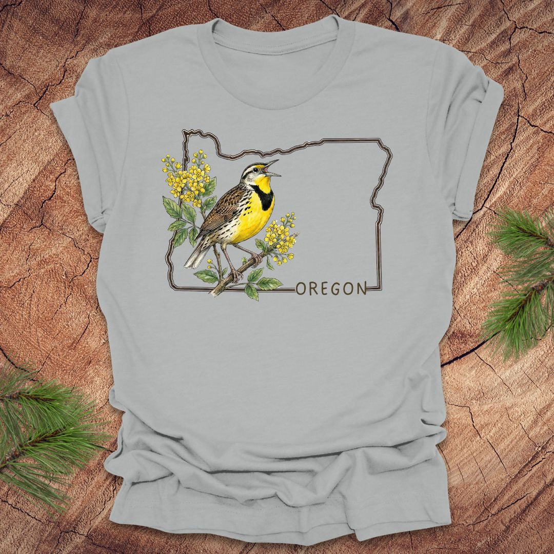 Silver t-shirt with a bird and floral design on an outline of Oregon, placed on a wooden surface.